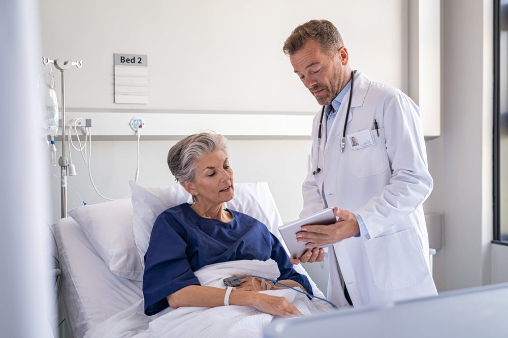 Serious doctor showing on digital tablet the medical report and health results to senior patient. Old woman sitting on hospital bed listening surgeon while explaining surgery procedure. 