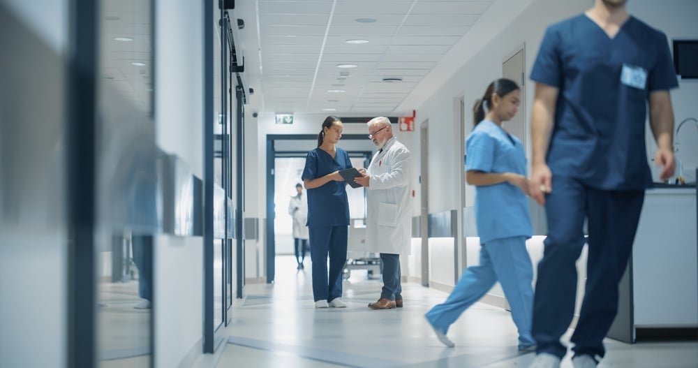 Portrait of a Young Caucasian Female Nurse and a Senior Male Doctor Discussing Patient Records in a Hospital Hallway on a Tablet Computer, Highlighting Efficient Healthcare Communication and Teamwork