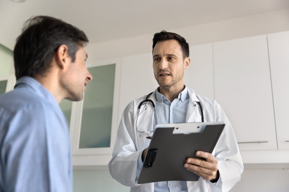 Serious general practitioner, cardiologist man talking to patient in clinic office, asking man about complaints, writing notes in paper records, giving consultation, prescription, recommendation